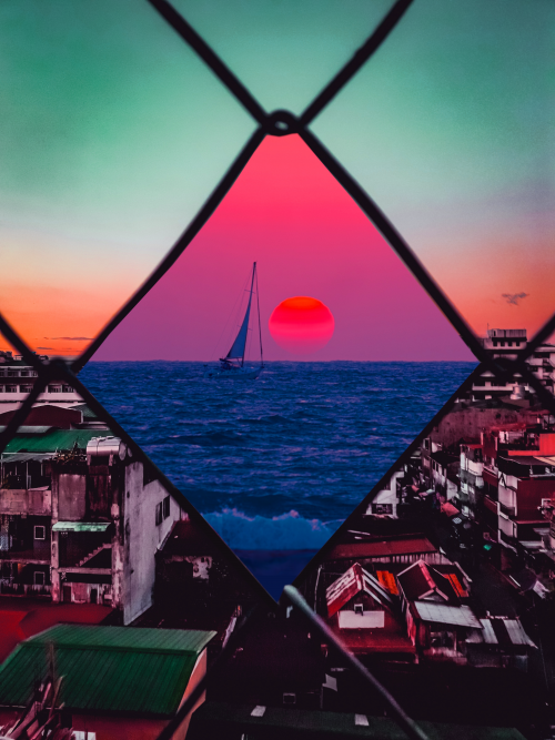 Image of a chain linked fence, the foreground slum-like while the center of the chain shows a beautiful sail boat coasting in the sunset